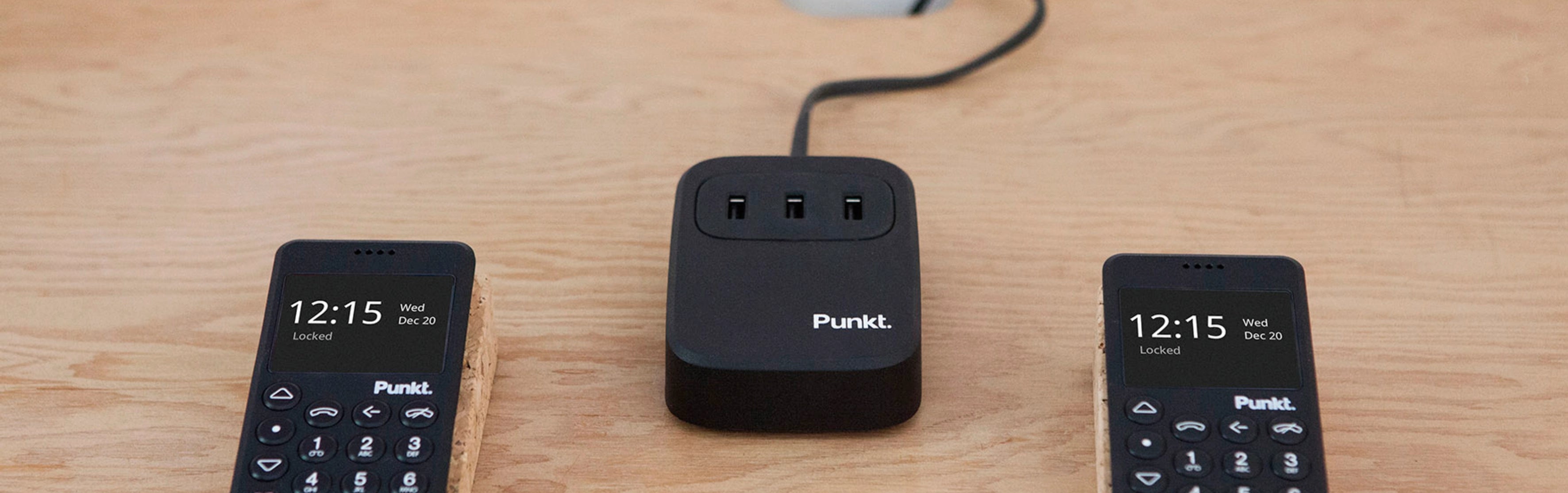 Punkt UC01 USB charger placed on a wooden desk, flanked by two MP02 phones, showing a clean and efficient charging setup.