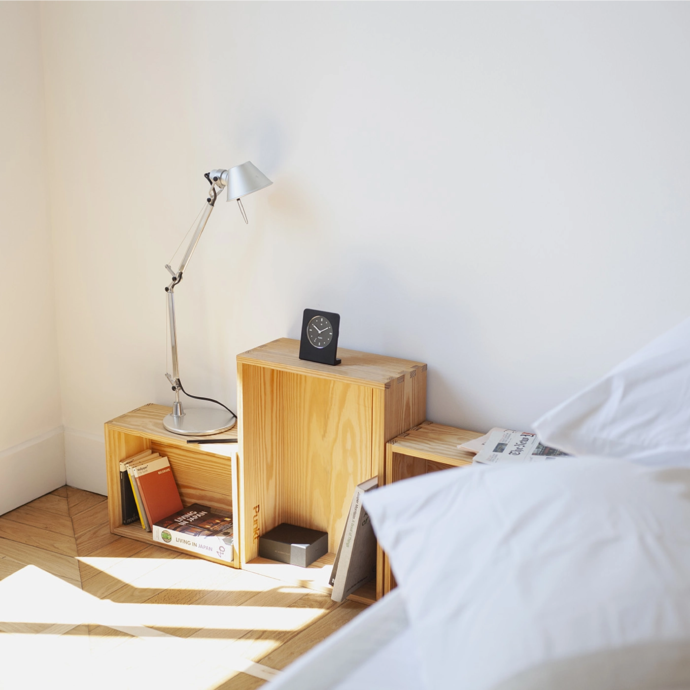 Black minimalist alarm clock by Punkt. placed on stacked wooden crates used as a bedside table, next to a silver desk lamp, books, and a neatly made bed with white sheets.