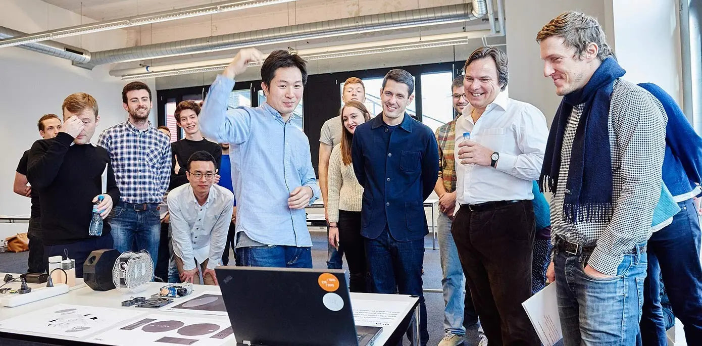 “A group of people standing and smiling around a table covered with electronics and prototypes during a lively workshop or demonstration.”