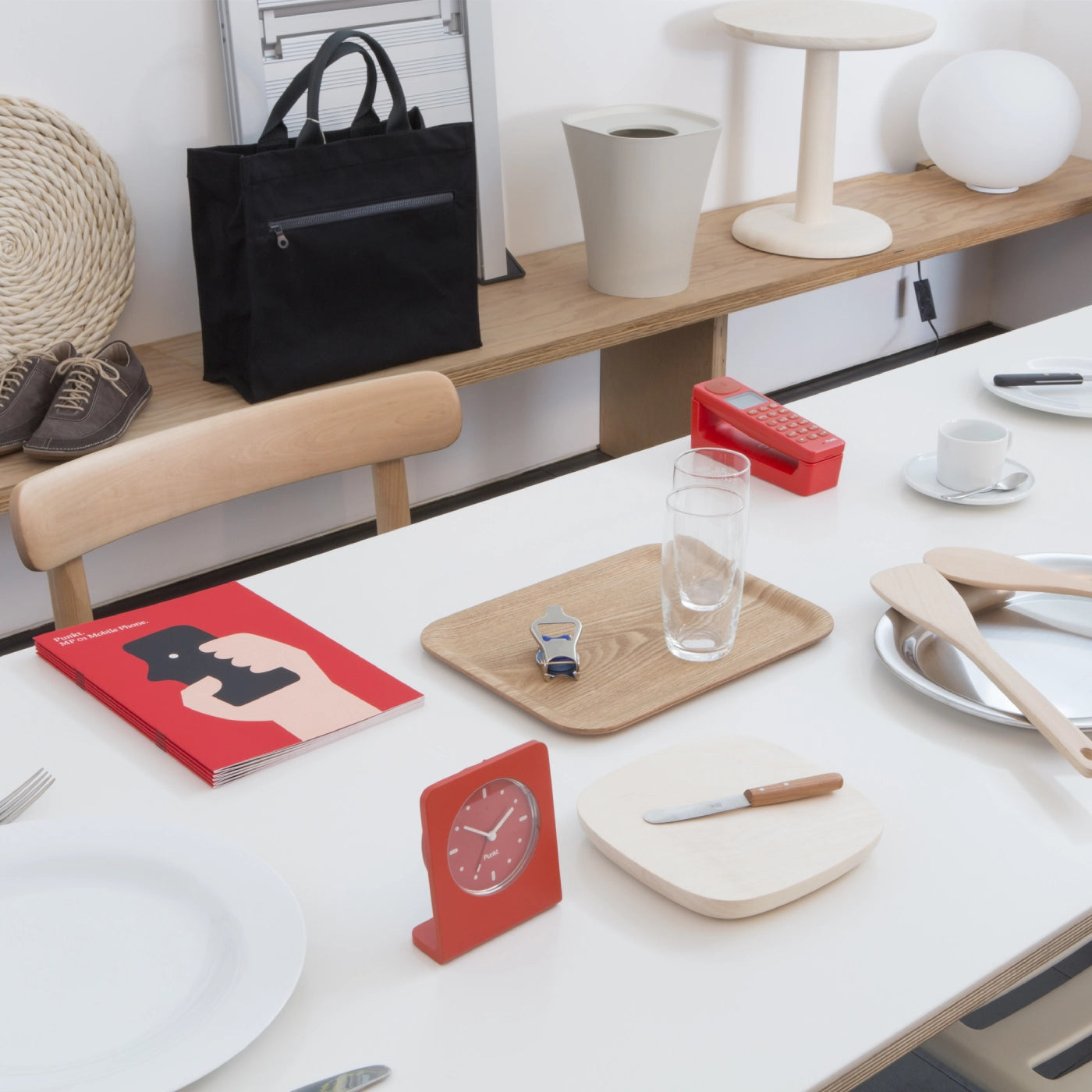 A white table set with plates, glasses, cutlery, and minimalist objects including a red Punkt. clock and a red Punkt. phone. The scene is bright and modern, featuring wooden elements and a stack of red booklets with a hand holding a phone illustrated on the cover.