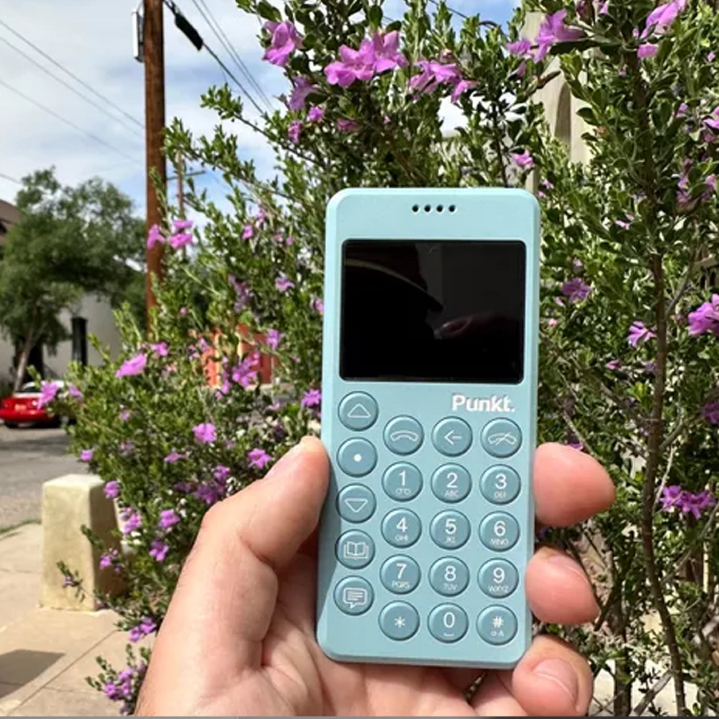 A hand holding the light blue Punkt. MP02 minimalist phone outdoors, with purple flowers and greenery in the blurred background.