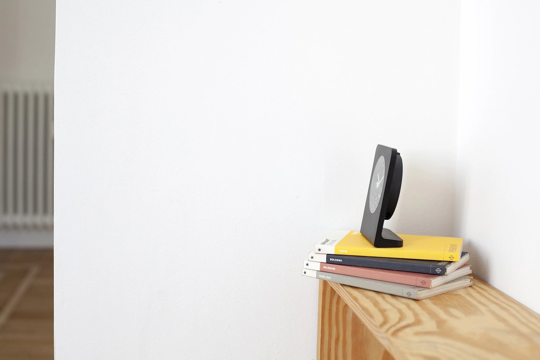 Side view of the Punkt AC02 clock in black, resting on a stack of travel guides, against a clean white wall.