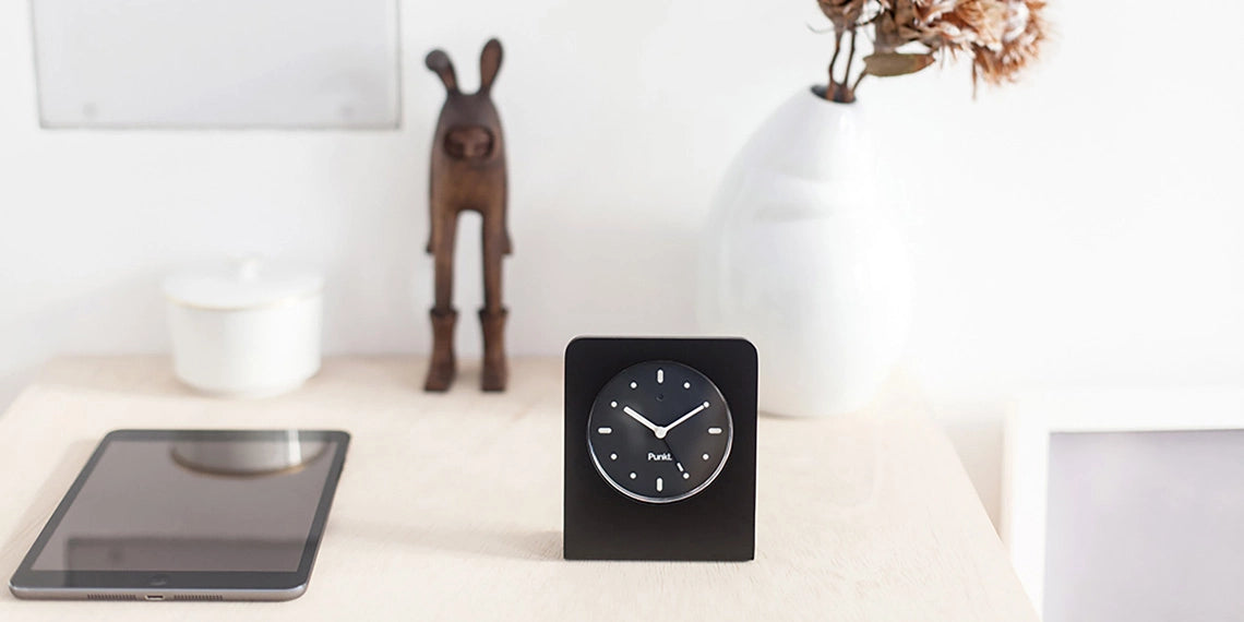 Punkt AC02 black clock on table – Black AC02 alarm clock standing on a wooden tabletop, clock face visible, with a phone in background slightly out of focus.