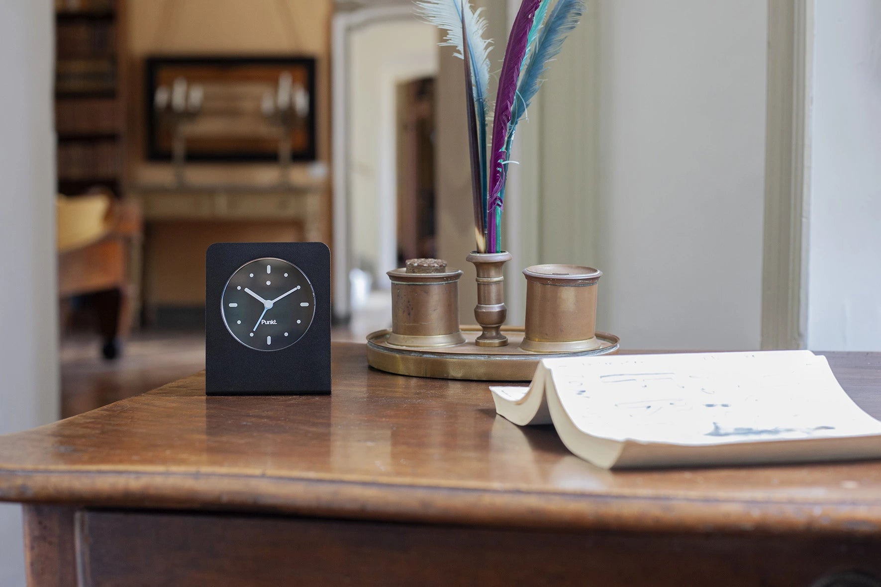 Punkt AC02 with pale blue second hand next to colorful feathers and brass inkwells, evoking a refined intellectual workspace.