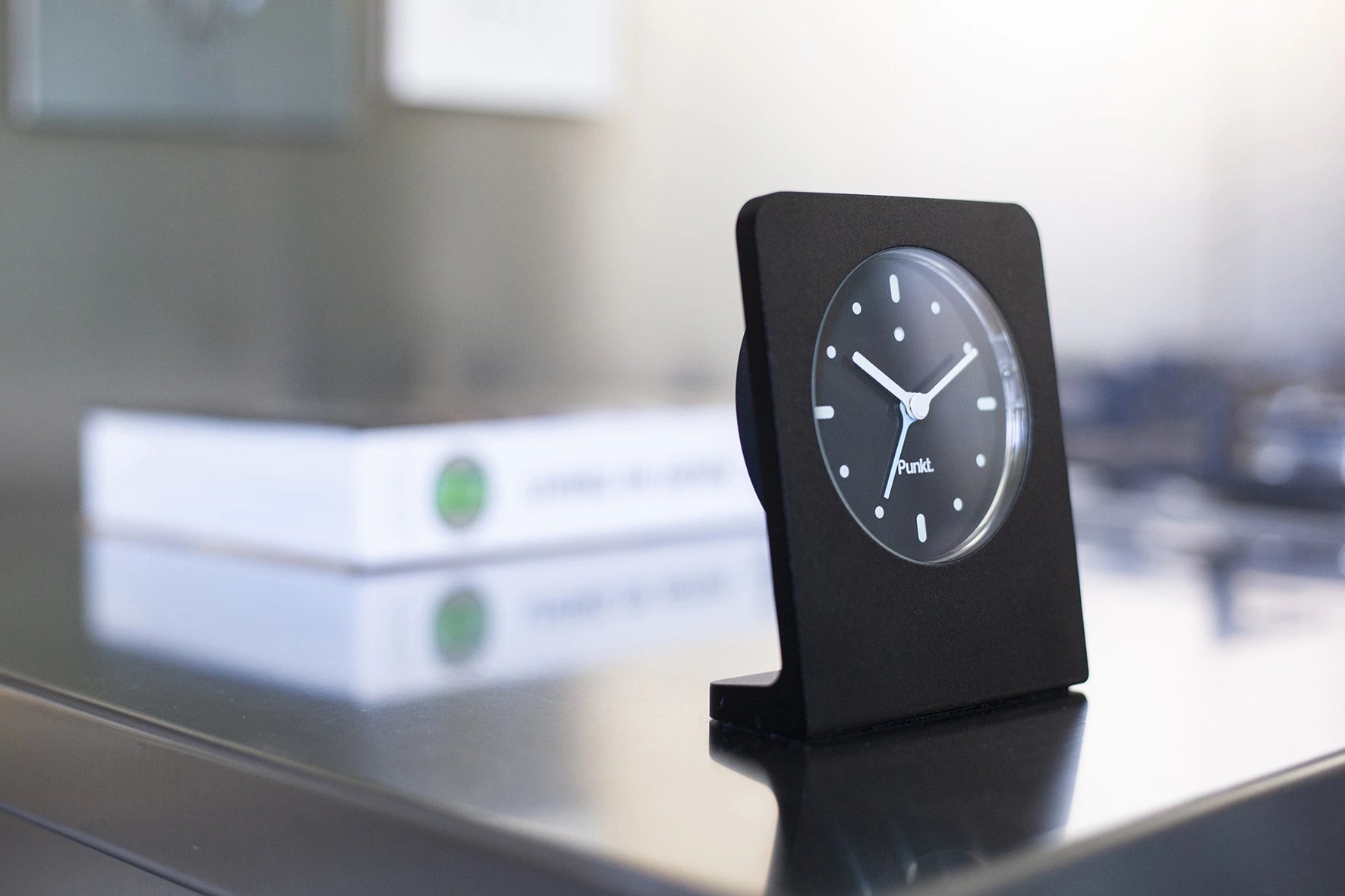 Punkt AC02 with blue second hand on a reflective stainless steel surface, with a design book in the background.