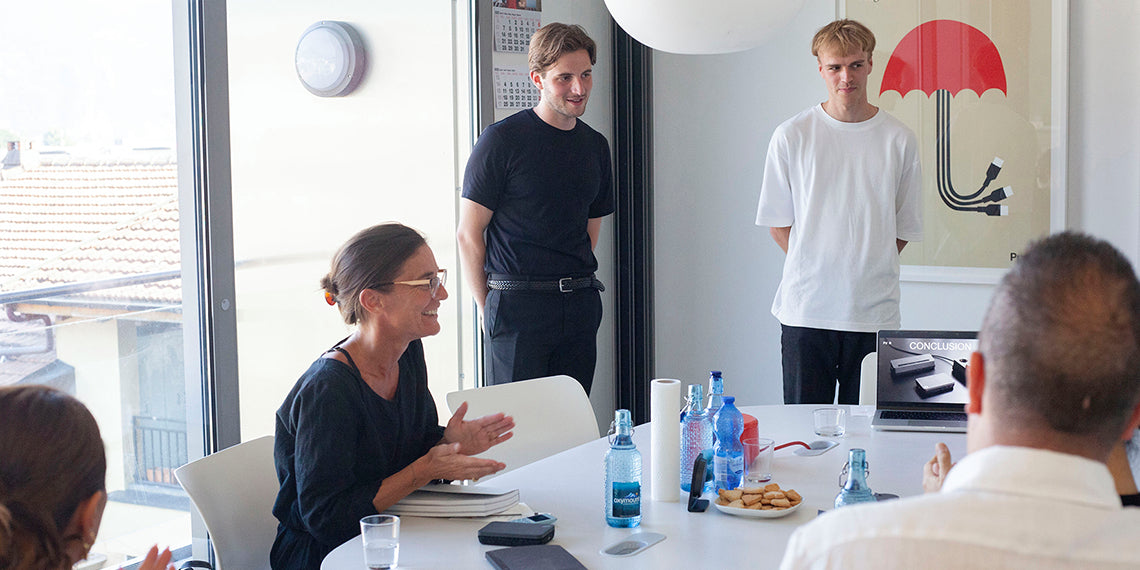 “People in a meeting room smiling as two men stand near a laptop showing a ‘Conclusion’ slide, with water bottles and cookies on the table.”