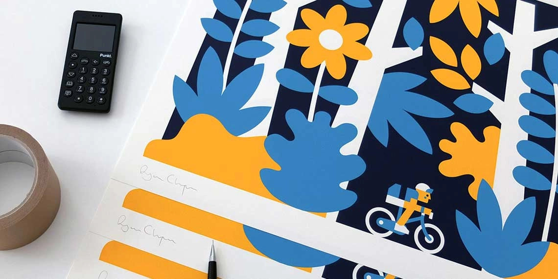 A black Punkt. phone sits on a white desk beside a roll of tape, a black pen, and a signed art print featuring a colorful, stylized forest scene in yellow, blue, and white with a character riding a bicycle through the trees.