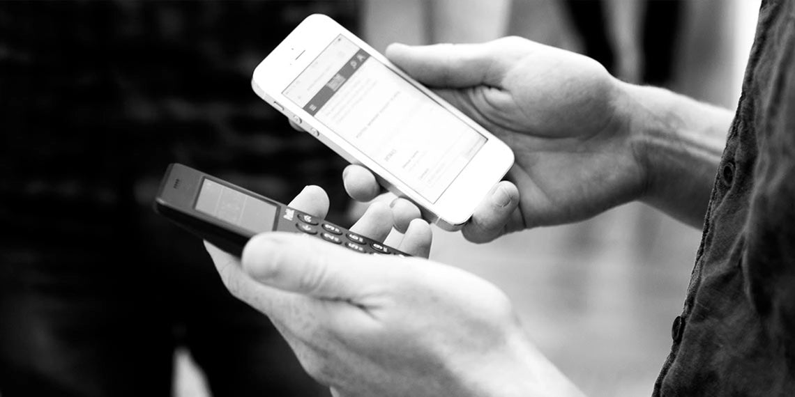 In a black-and-white photo, a person holds a Punkt. minimalist phone in one hand and a smartphone in the other, visually contrasting digital simplicity with a connected device.