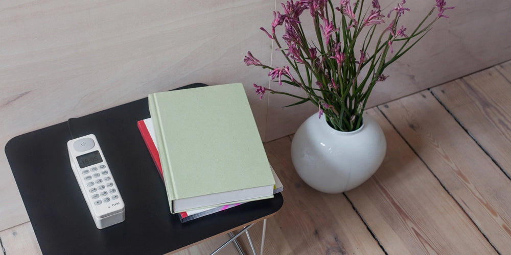 White Punkt. DP01 cordless phone on a black side table, with a stack of books and a white vase with pink flowers in a cozy, minimalist interior.