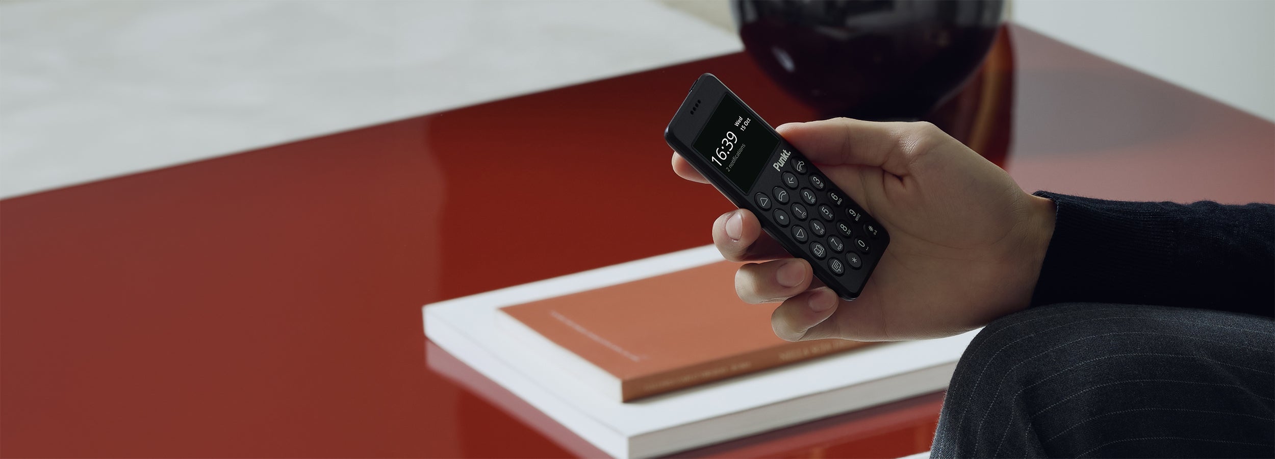 A close-up of a hand holding a black compact phone showing the time on its screen, placed near a red table with an orange book on it.