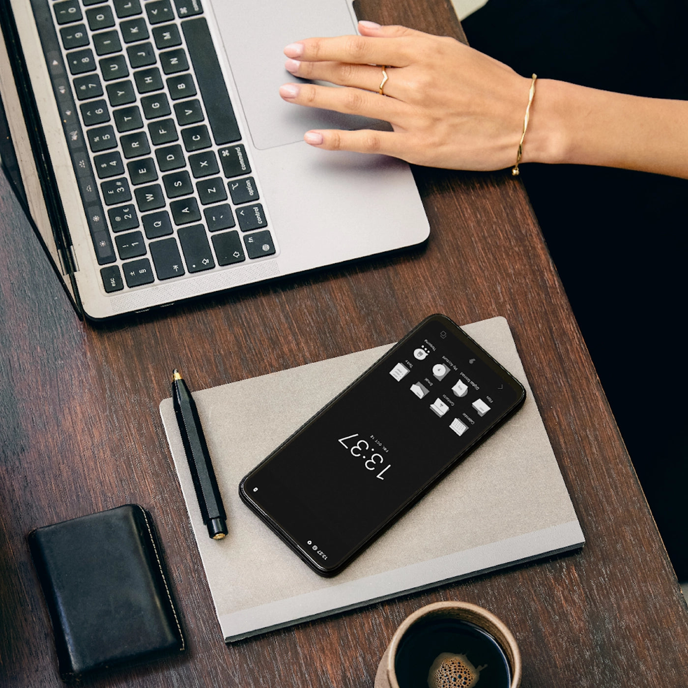 Overhead view of a minimalist workspace with a laptop, a notebook, a pen, a wallet, a cup of coffee, and a Punkt MC02 smartphone displaying a monochrome home screen and clock.