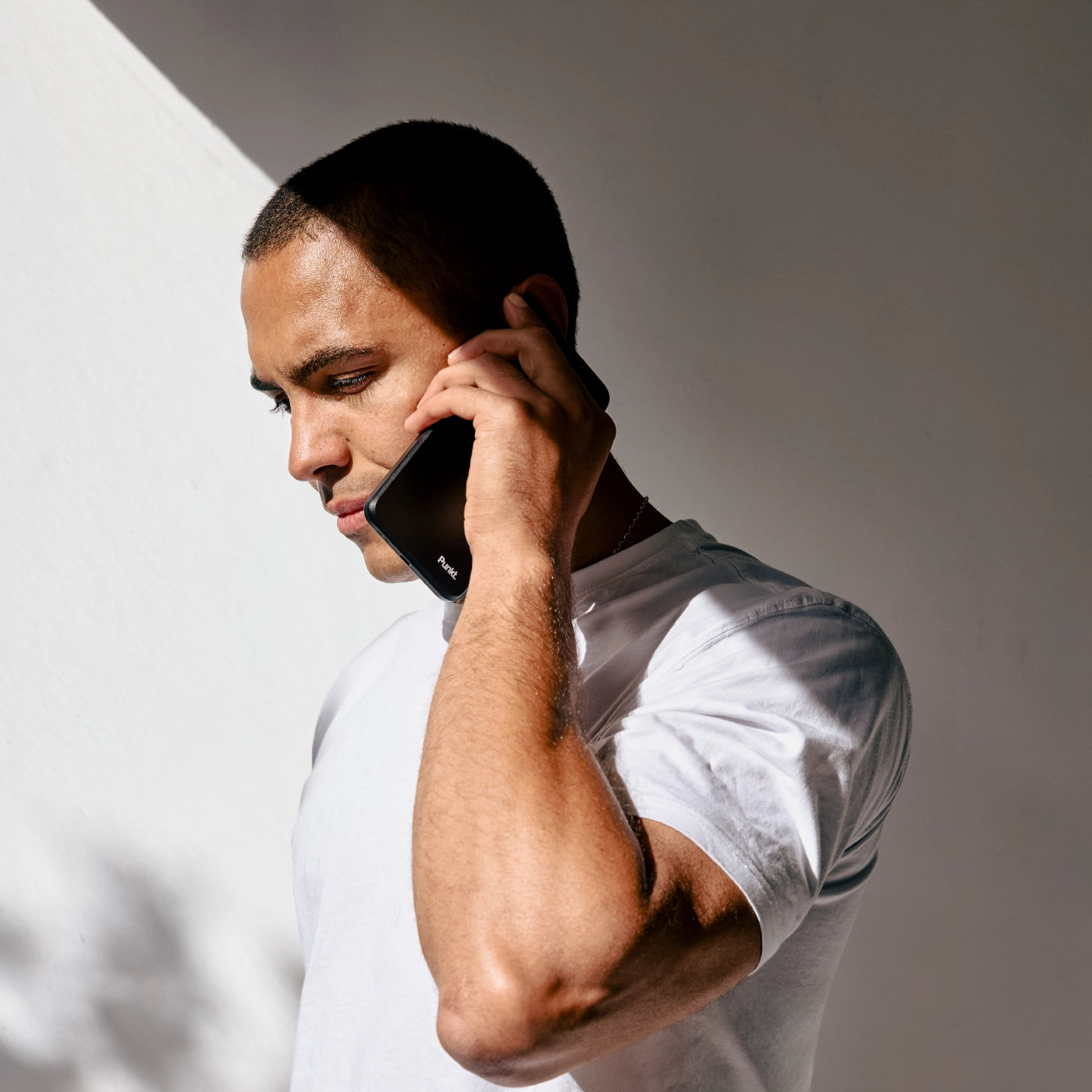 Man in white t-shirt talking on black phone against gray background.