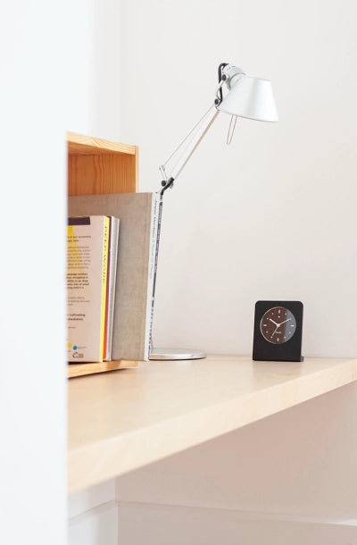 Black Punkt. AC01 alarm clock placed on a light wood desk next to a silver desk lamp and a bookshelf, in a clean minimalist workspace.