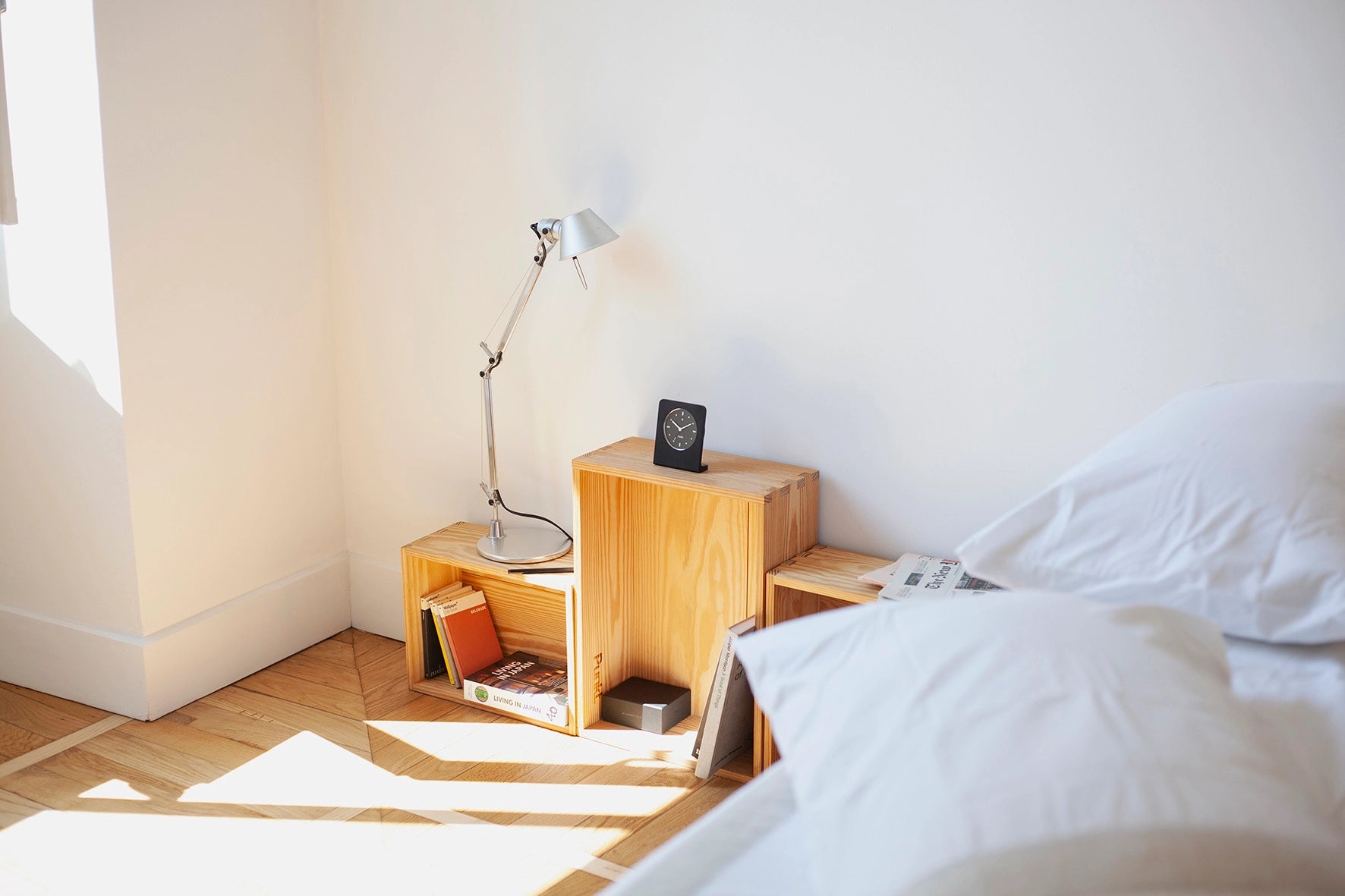 Punkt. clock placed on a stack of wooden crates used as a bedside table, in a sunlit minimalist bedroom.