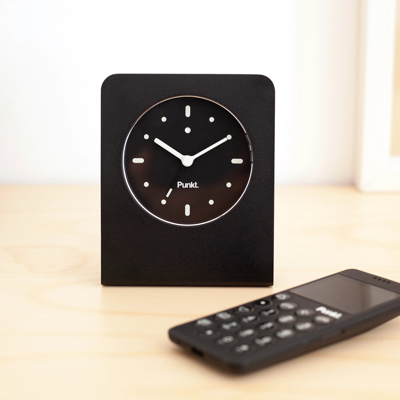 Minimalist black Punkt. AC02 alarm clock and MP02 mobile phone placed on a light wooden surface in a clean, modern interior.