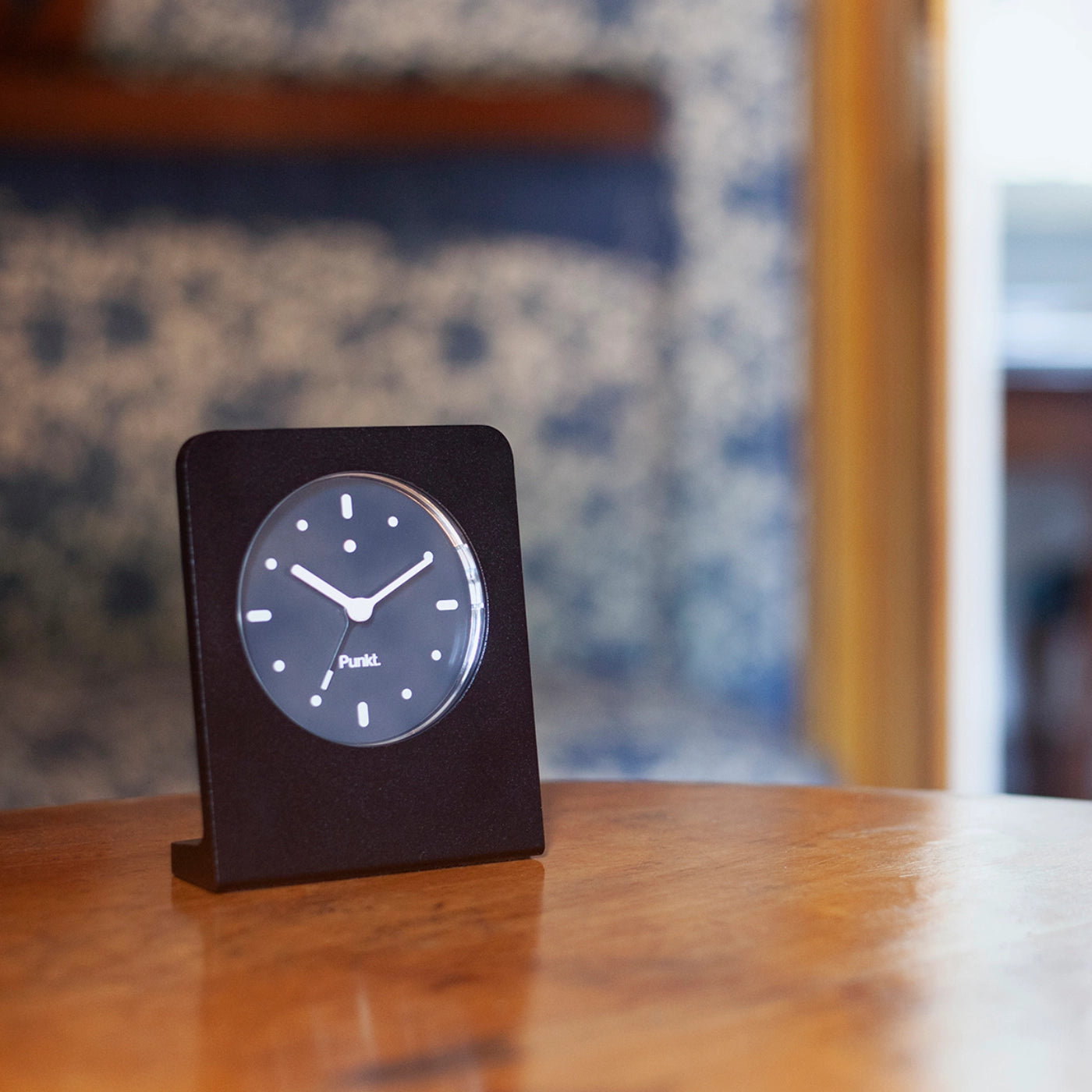 Black Punkt. AC02 alarm clock on a wooden table in a warmly lit room with a patterned blue wall in the background.