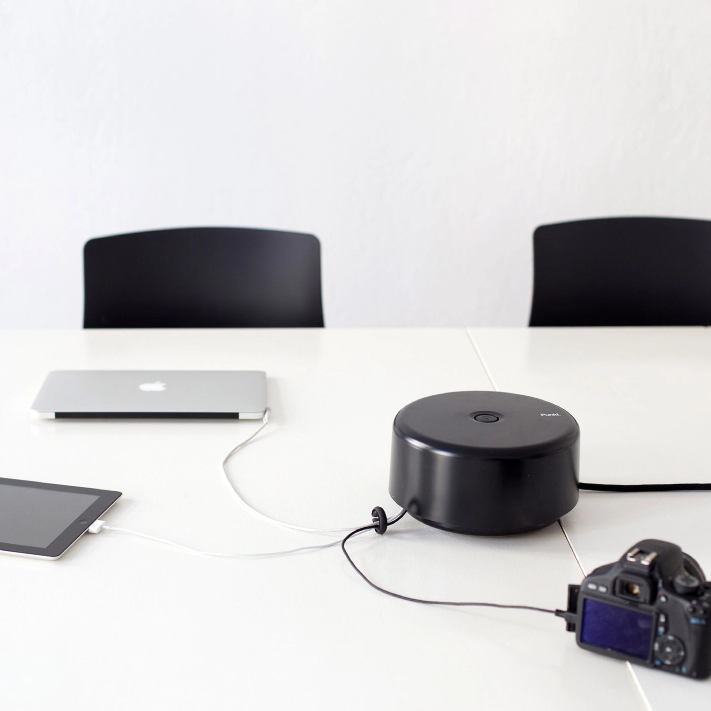 Punkt. ES01 extension socket in black, placed on a white table powering a laptop, tablet, and camera in a minimal office setup.