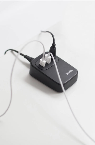 A black Punkt. UC01 USB charging station with minimalist design, plugged in and actively charging multiple devices with black and white cables, resting on a sleek white surface.