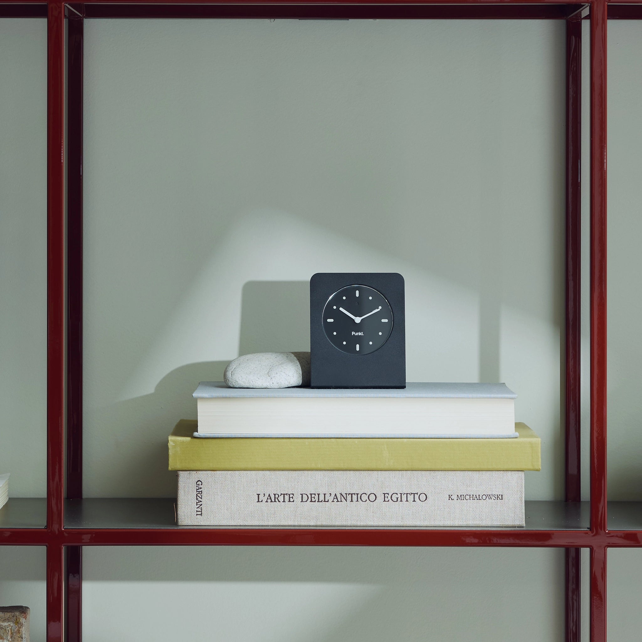 A small black desk clock placed on top of stacked books on a red metal shelf, with a smooth gray wall in the background.