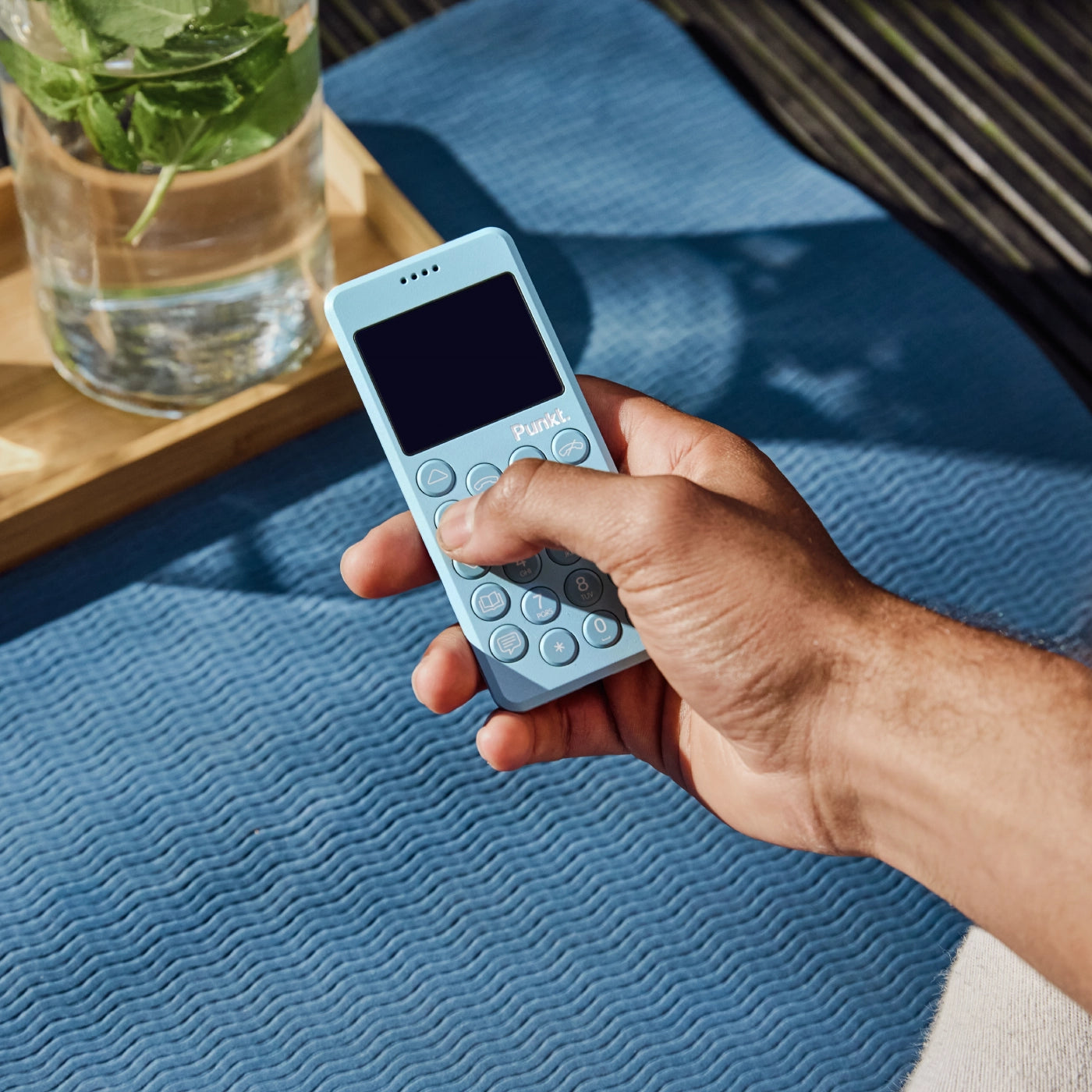 A hand holding the blue Punkt. MP02 over a wavy-textured blue mat, next to a glass jug with mint leaves.