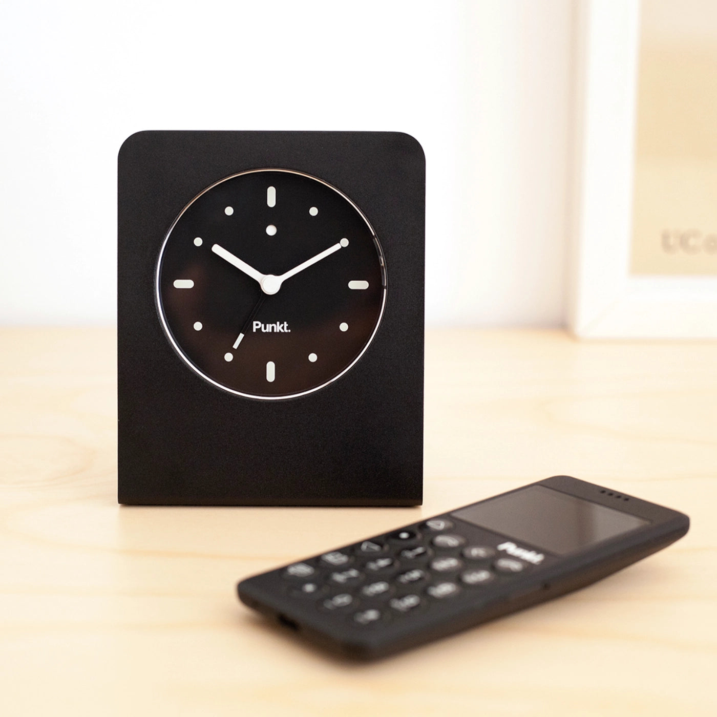 Punkt AC02 alarm clock with black second hand placed beside the MP02 minimalist mobile phone in black, styled on a light wooden surface.