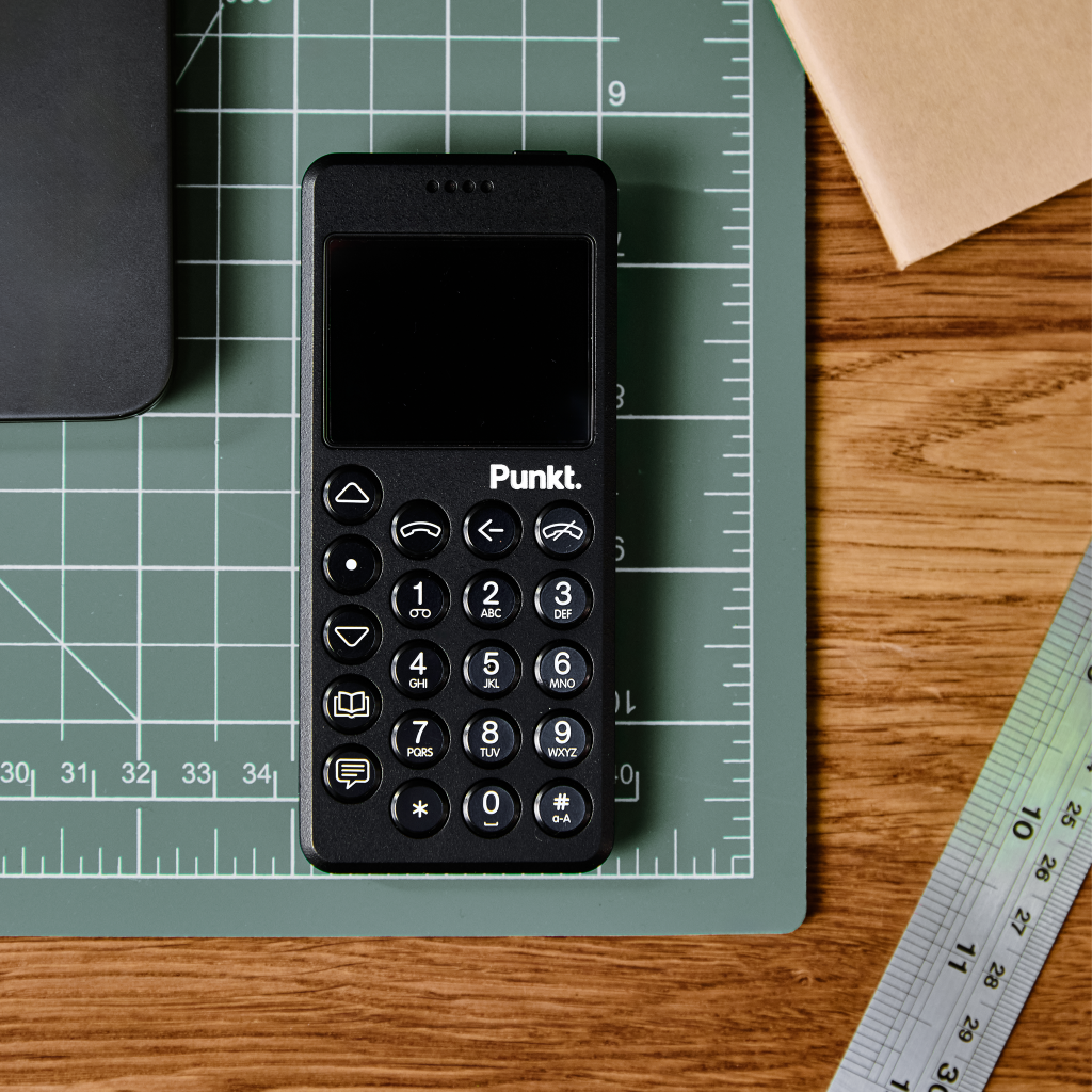 Black Punkt. MP02 phone resting on a green cutting mat beside a wooden desk and ruler.