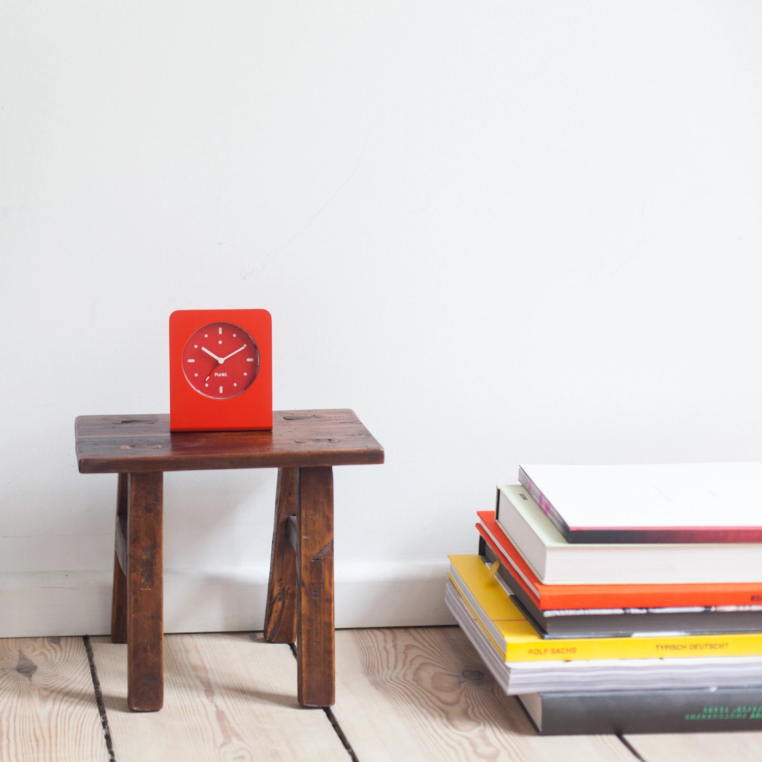 Punkt. AC01 analog alarm clock in red, displayed on a rustic wooden stool next to a stack of books against a white wall.