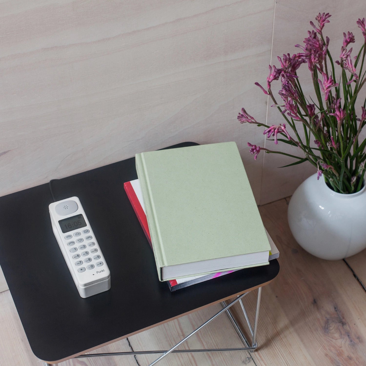 Punkt. DP01 cordless phone in white, placed on a black side table with stacked books and a white vase with pink flowers in a cozy interior setting.