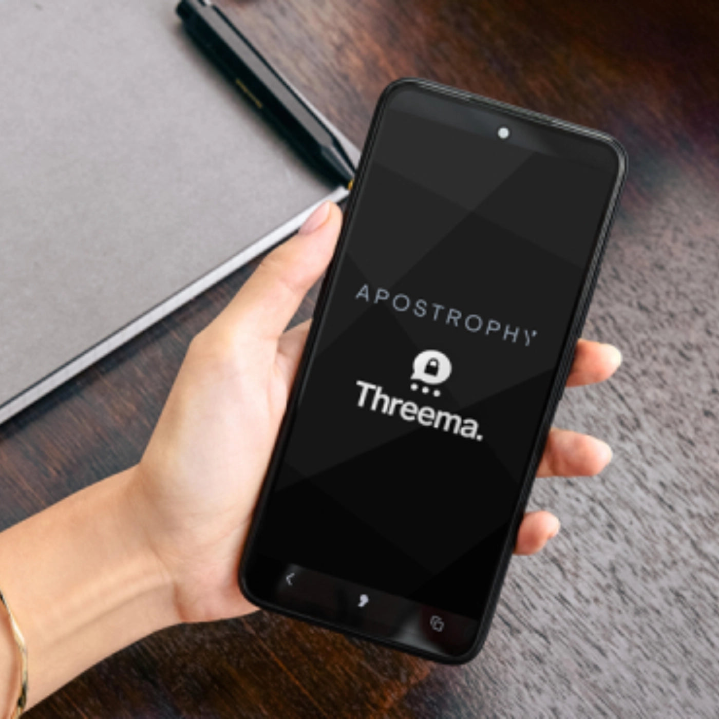 Hand holding a smartphone displaying the Apostrophy and Threema logos on screen, placed above a dark wooden desk.