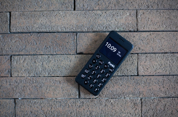 Top view of a black Punkt. MP02 minimalist phone resting on a textured brick wall background, screen displaying the time 10:09.