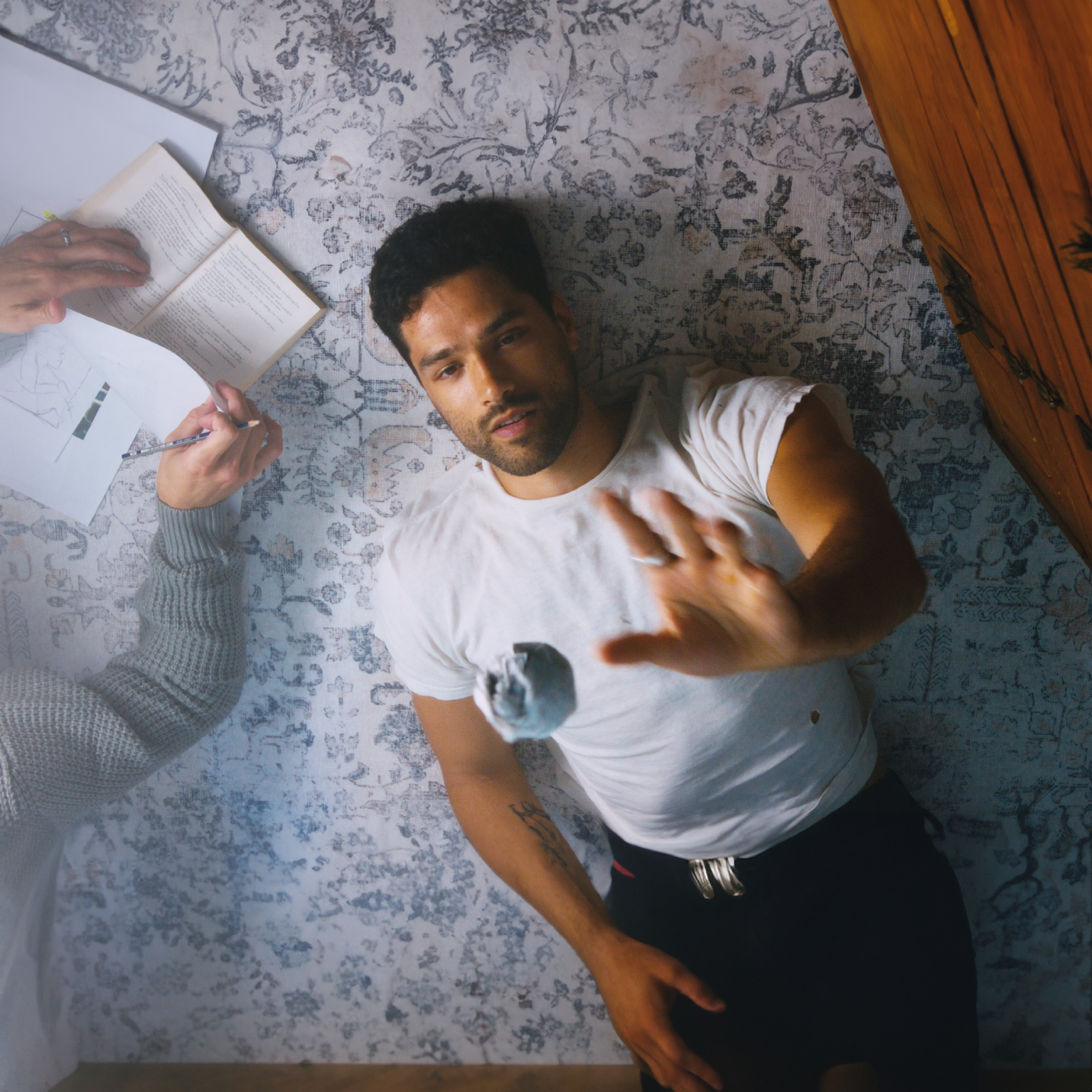 A young man in a white t-shirt lies on a patterned rug, looking up while gently tossing a small fabric object in the air. Next to him, someone reads a book and writes on papers spread out.
