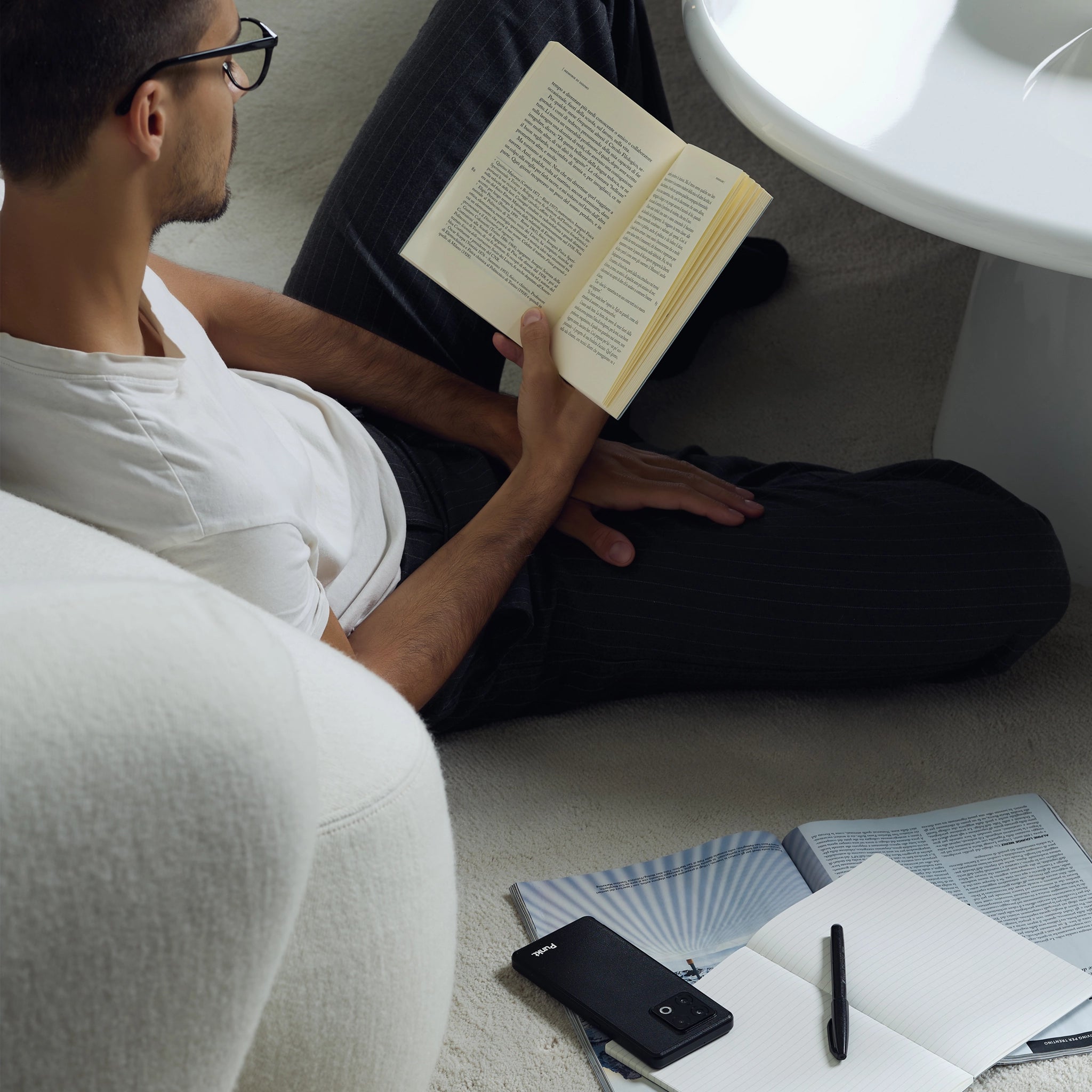 Man reading a book on the floor with a phone, pen, and papers nearby.