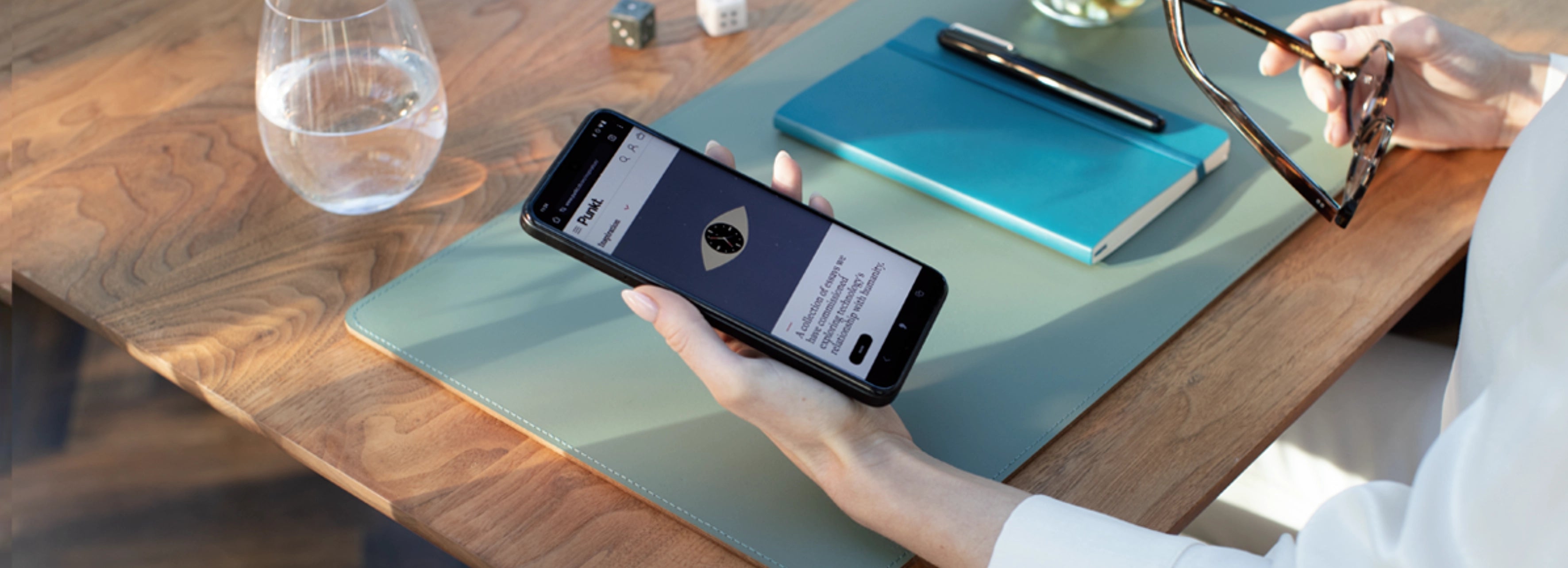 Person holding smartphone showing an app interface, sitting at wooden desk with notebook, glass of water, and eyeglasses.