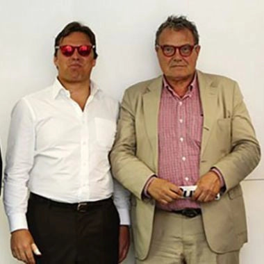 A cropped version of the previous image showing two men in red sunglasses — one in a white shirt and the other in a tan suit and red checkered shirt — standing side by side against a white wall.