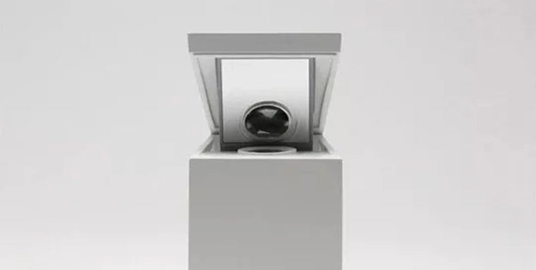 A minimalistic, modern cube-like object with a lid partially open reveals a single glossy black orb inside, placed in the center of a white gallery or showroom with a neutral backdrop.