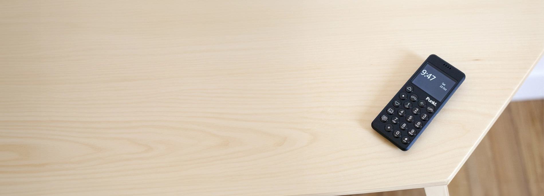 A black Punkt minimalist phone with physical buttons and digital display showing the time 9:47, placed on a light wooden table.
