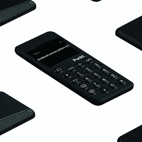 Multiple black Punkt. MP02 phones on dark background – Angled view of several black minimalist phones scattered across a dark surface, one screen showing “Archived conversations (1)”.