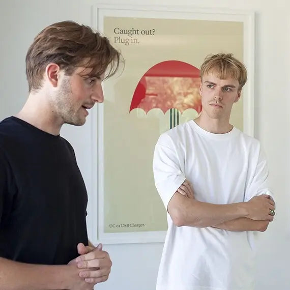 “Two young men standing in front of a poster with a red umbrella illustration, one speaking while the other listens with arms folded.”