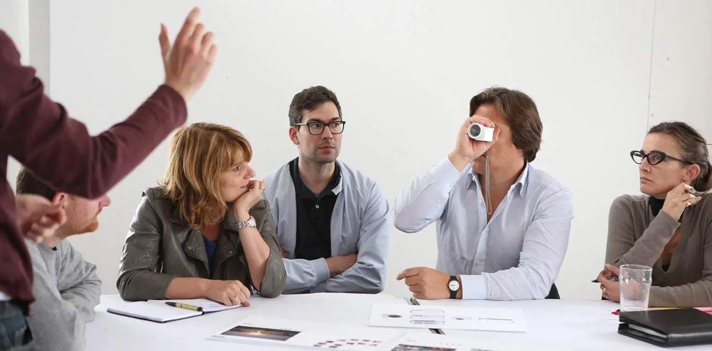“Group of people sitting around a meeting table while one man peers through a small cylindrical device, with papers and glasses of water on the table.”