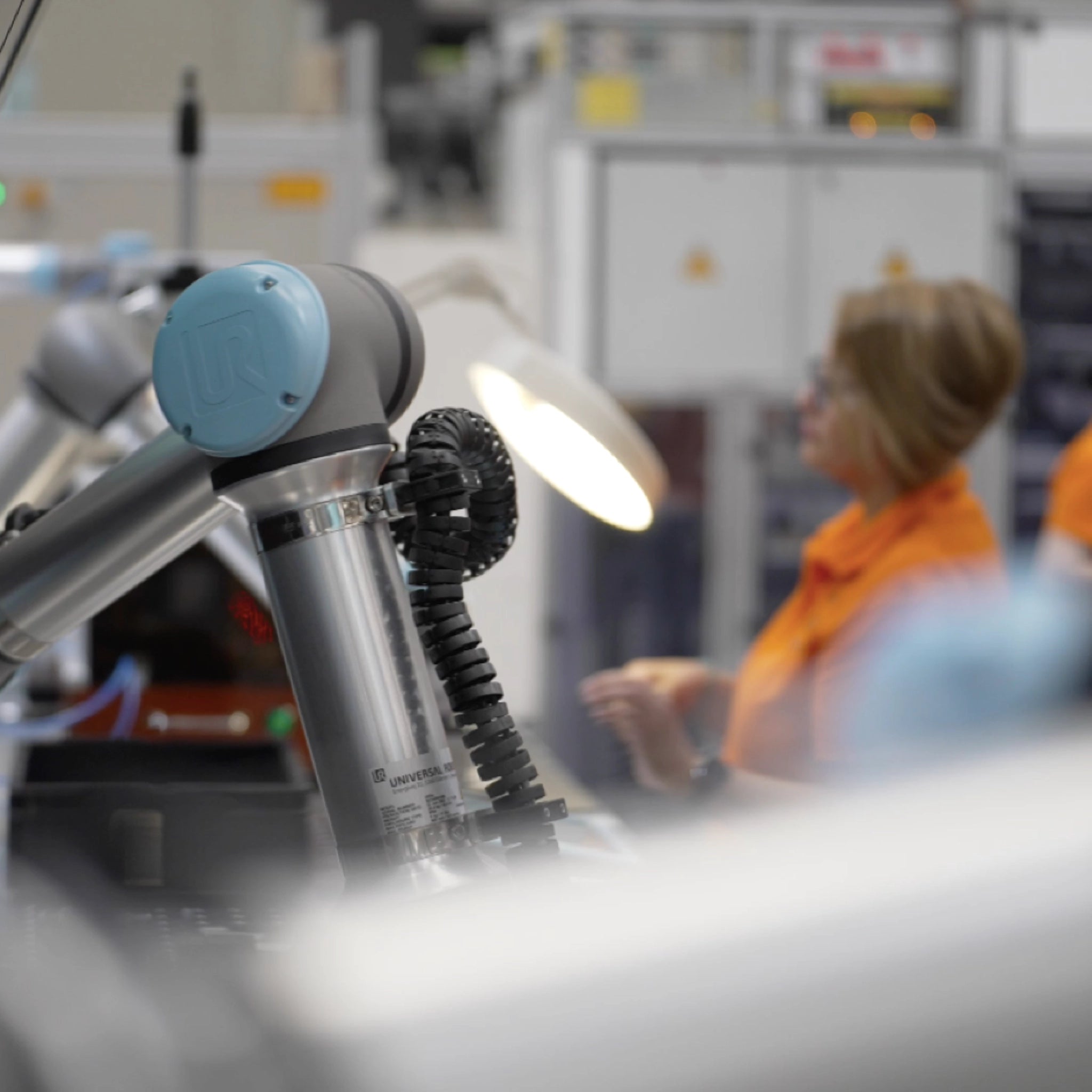 Robotic arm in a factory setting with a person in an orange jacket in the background.