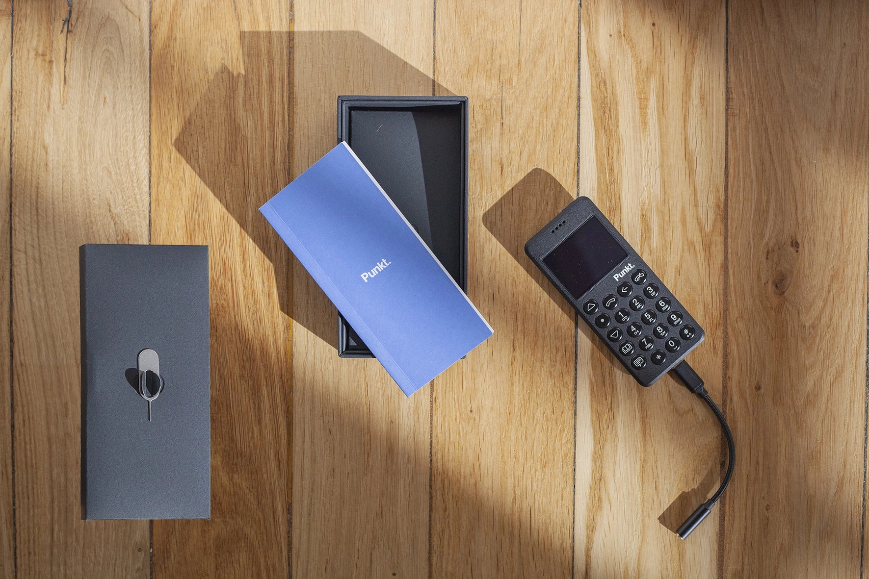 Flat lay of a black Punkt. MP02 phone partially charged, its packaging, and instruction booklet on a wooden surface.