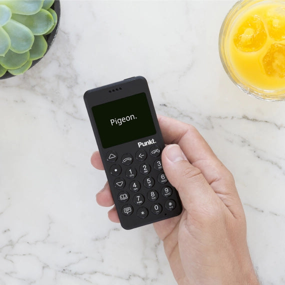A hand holding a black Punkt. phone displaying the word "Pigeon." on the screen, placed on a white marble surface next to a glass of orange juice and a green succulent plant.