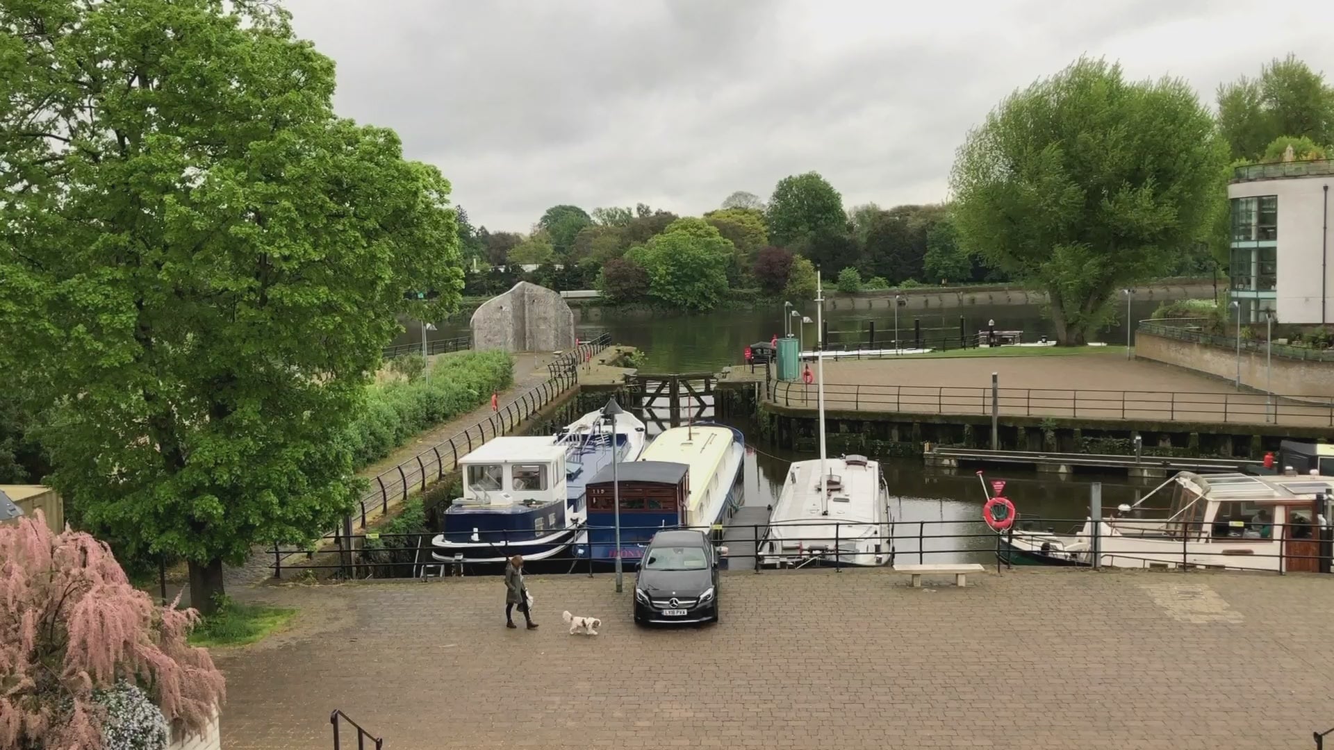 Video thumbnail showing a riverside view with moored boats, a car, and a person walking a dog near green trees and calm water.