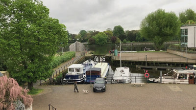 Video thumbnail showing a riverside view with moored boats, a car, and a person walking a dog near green trees and calm water.