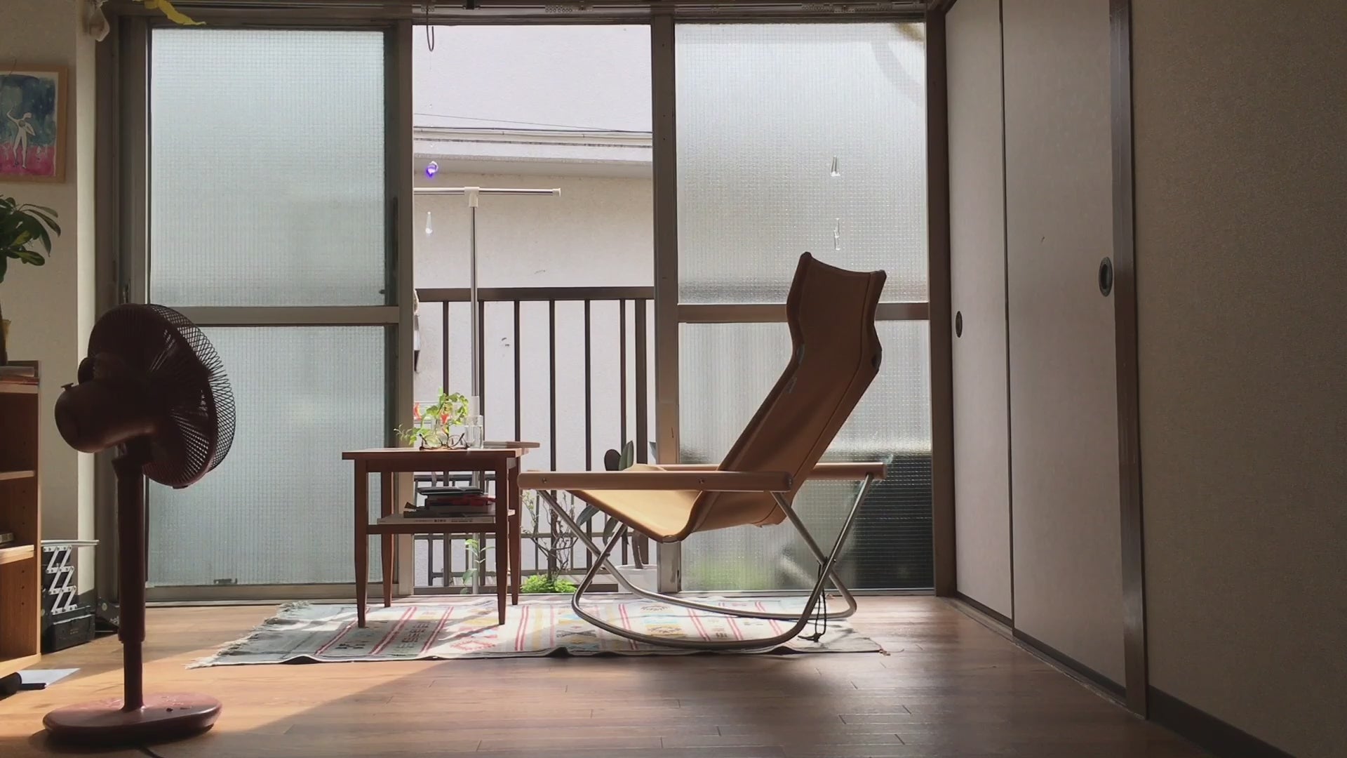 Video thumbnail showing a peaceful interior with a modern rocking chair, wooden table, potted plants, and a standing fan, softly lit by natural light from large frosted windows.