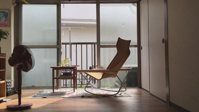 Video thumbnail showing a peaceful interior with a modern rocking chair, wooden table, potted plants, and a standing fan, softly lit by natural light from large frosted windows.