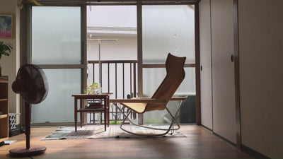Video thumbnail showing a peaceful interior with a modern rocking chair, wooden table, potted plants, and a standing fan, softly lit by natural light from large frosted windows.