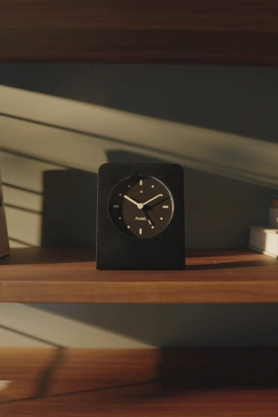 A cozy bedroom corner with warm light on wooden shelves and a minimalist Punkt. clock among books and decor.