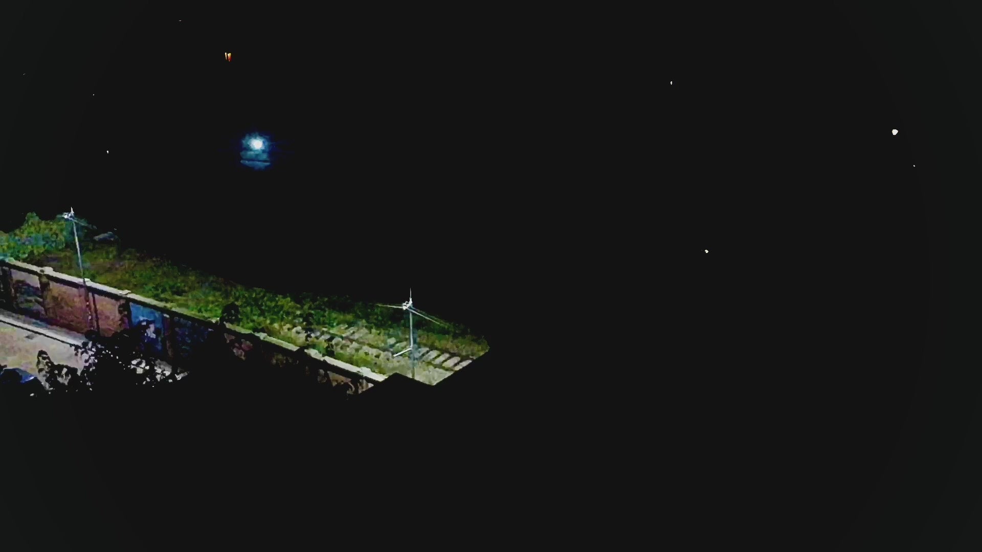 Video thumbnail showing a dark night scene with a small section of illuminated urban greenery, streetlights, and a fence, surrounded by deep black sky and scattered points of light.