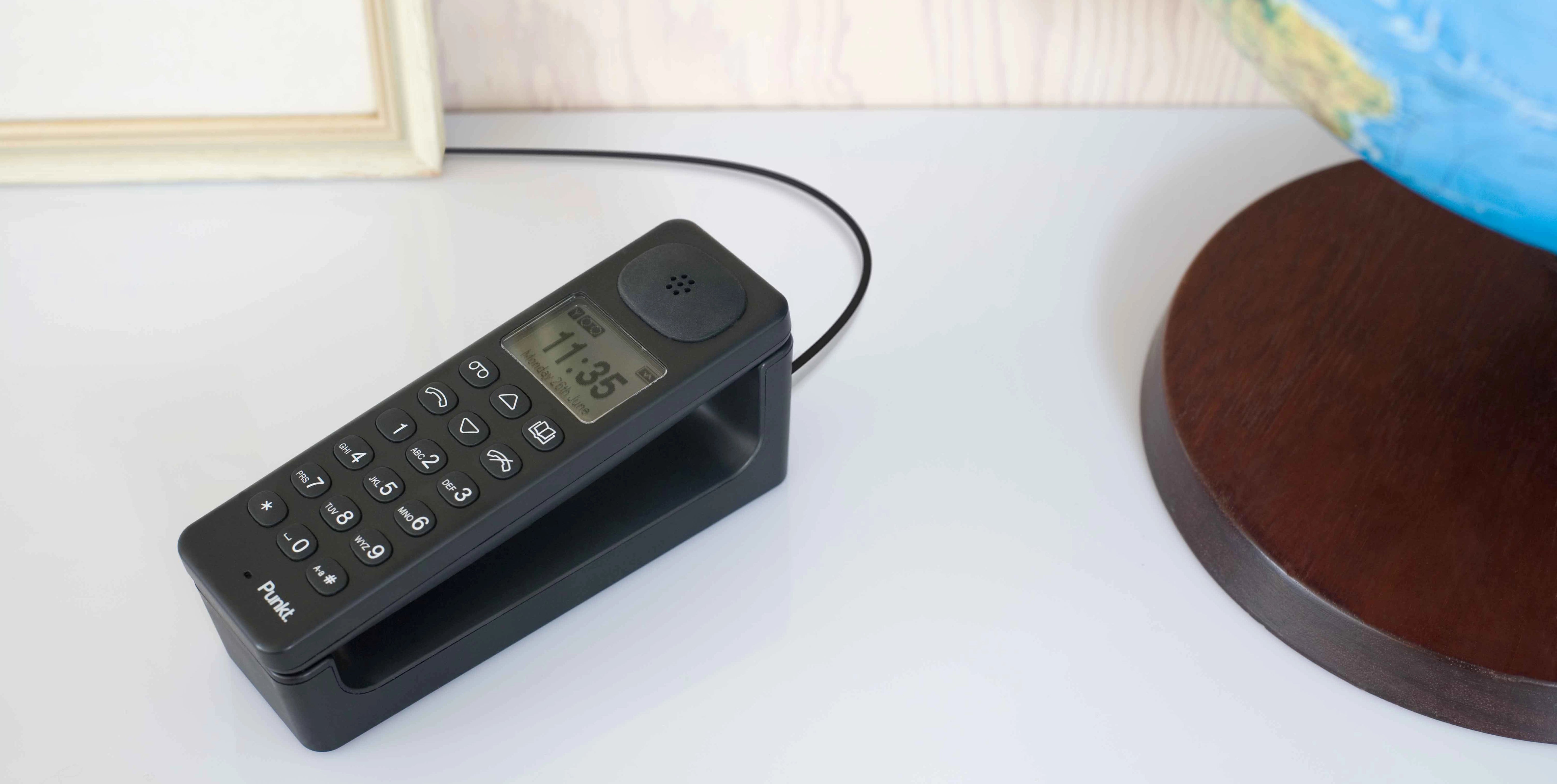 Punkt. DP01 cordless phone in black, placed on a white surface with a charging base, next to a globe and picture frame — minimal and functional desk setup.